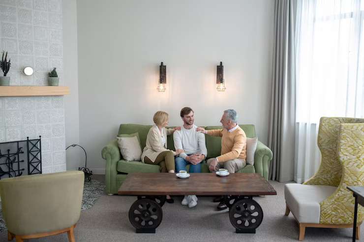 Adult son sitting on a sofa with his parents in a bright living room, talking and sharing coffee together at a coffee table.