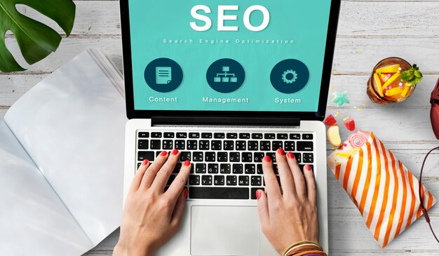Hands typing on laptop showing SEO screen with icons, desk setup with snacks, plant, and office items