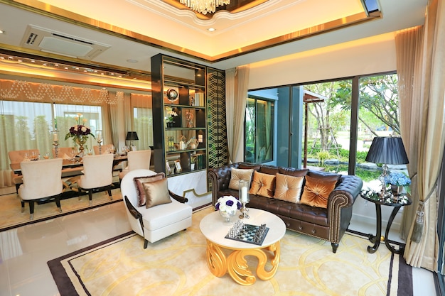 A luxurious living room with elegant furniture, a chandelier, large windows, and a dining area in the background.