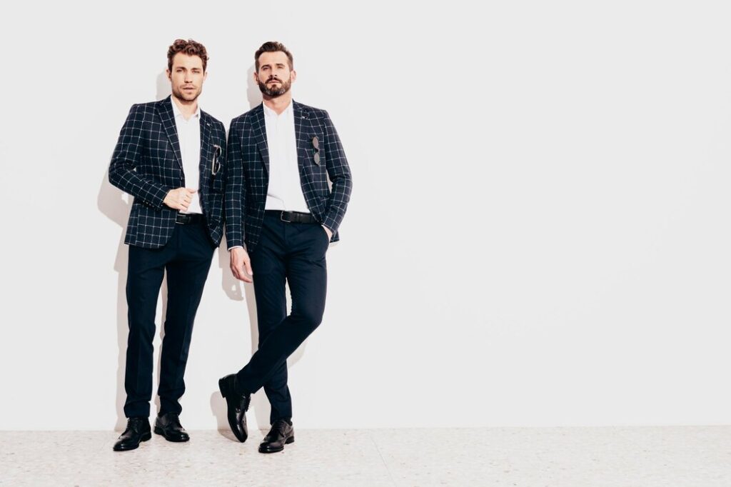 Two men in checkered blazers and dark pants posing against a plain white background.
