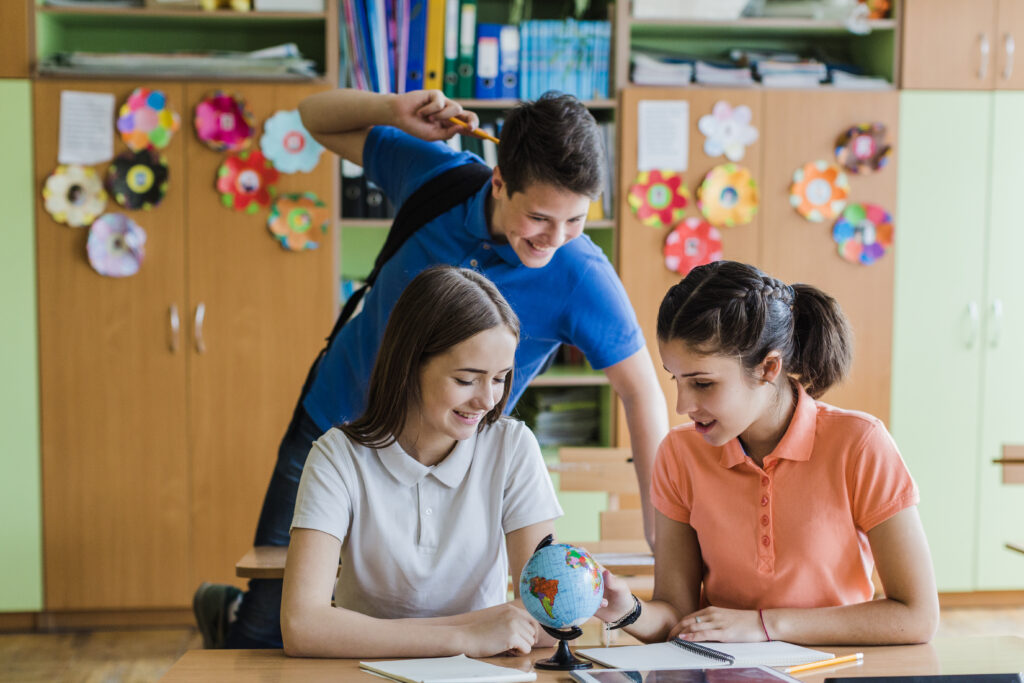 Two girls examine a small globe at a classroom desk while a boy playfully interacts behind them, surrounded by colorful decor.