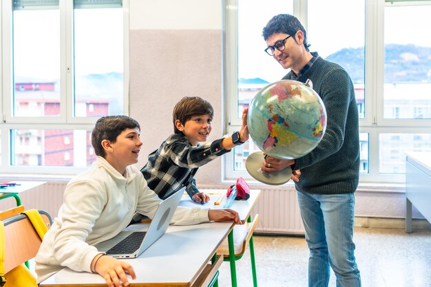 A teacher holds a globe while two young students at their desks engage excitedly, pointing and learning geography in class.