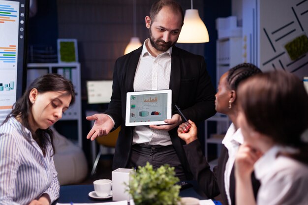 A businessman presenting data on a tablet to a group of colleagues during a meeting in a modern office.