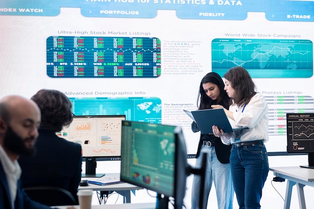Business professionals analyzing data on computer screens and a large digital display board in a modern office setting, with charts, stock market listings, and statistical information in the background.