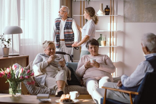 Elderly people enjoying tea together in a cozy living room, while a caregiver assists a man with a walker near the window.