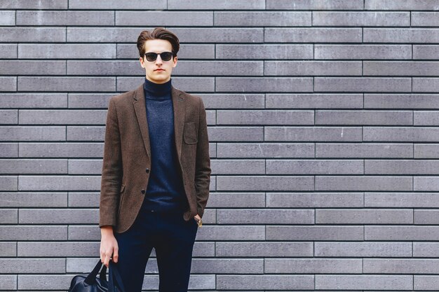 Stylish man in sunglasses wearing a brown blazer and dark turtleneck, holding a bag and standing against a gray brick wall.
