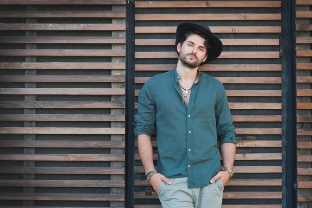 Man in a dark hat and green shirt leans against a wooden slatted wall with hands in pockets, giving a relaxed pose.
