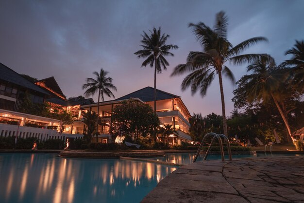 Luxury resort at dusk with palm trees, glowing lights, pool reflections, elegant architecture, and tropical surroundings.