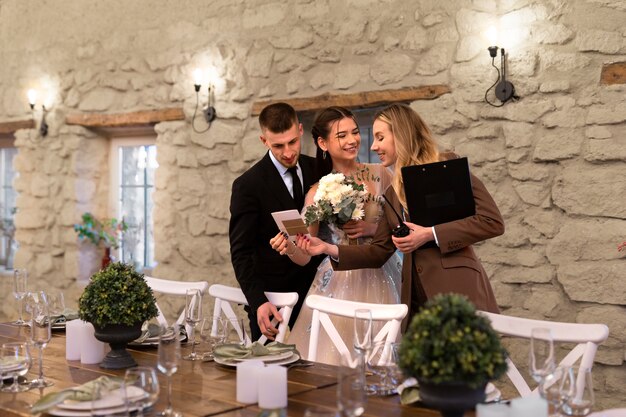 The wedding couple reviewing event details with the planner beside a decorated table inside a rustic celebration venue setting.