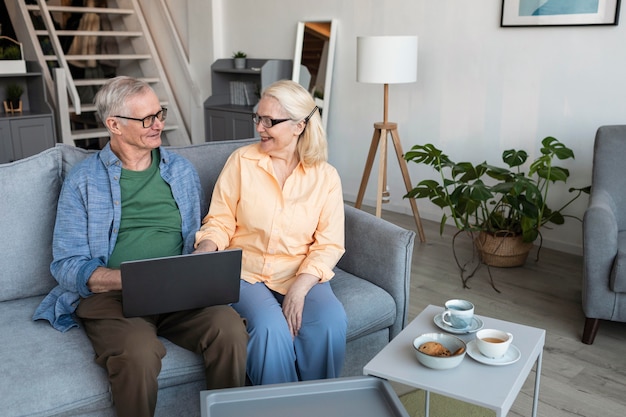 A senior couple sits on a sofa at home, smiling and chatting while using a laptop, with coffee cups on a nearby table.