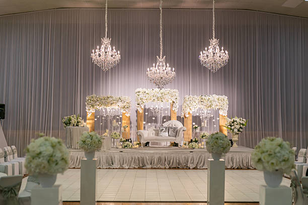 Elegant wedding stage with white floral arches, crystal chandeliers, soft drapes, and a decorated aisle in an indoor hall.